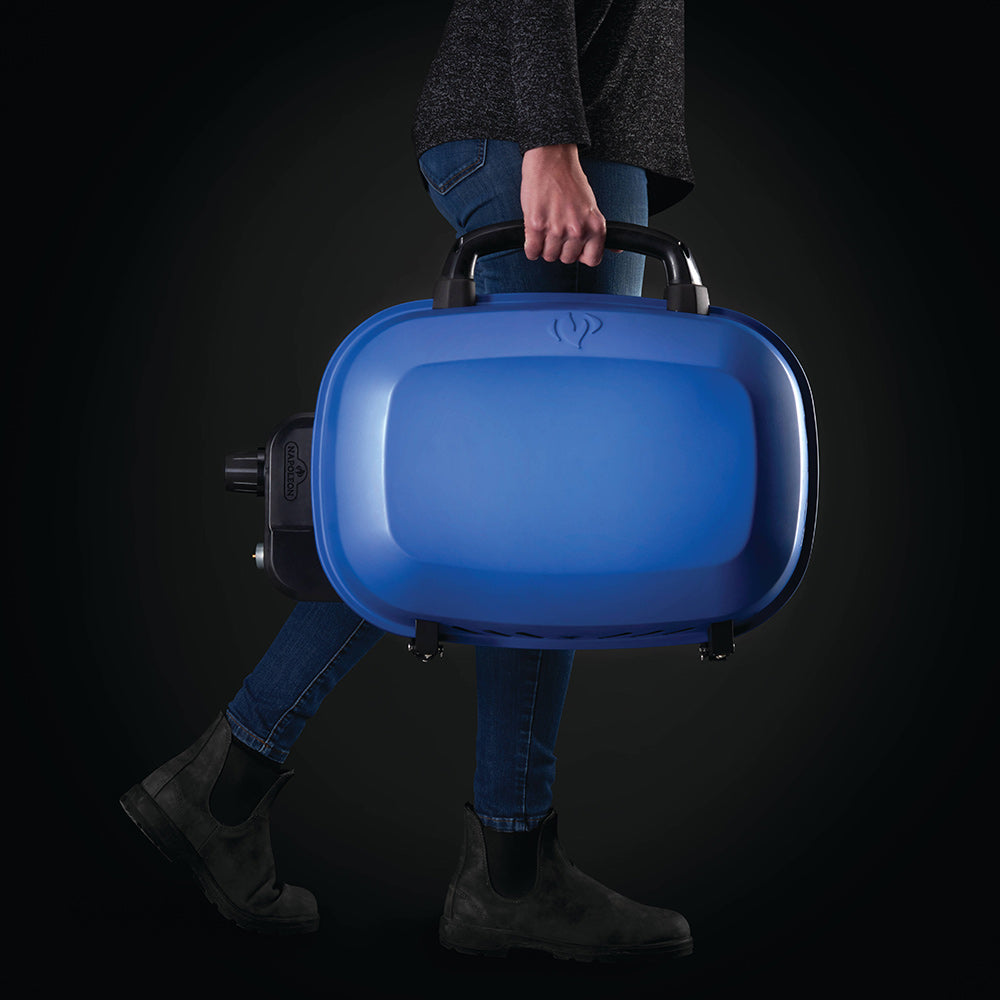 Blue portable tabletop gas grill, lid closed, carried by a person with black side handle
