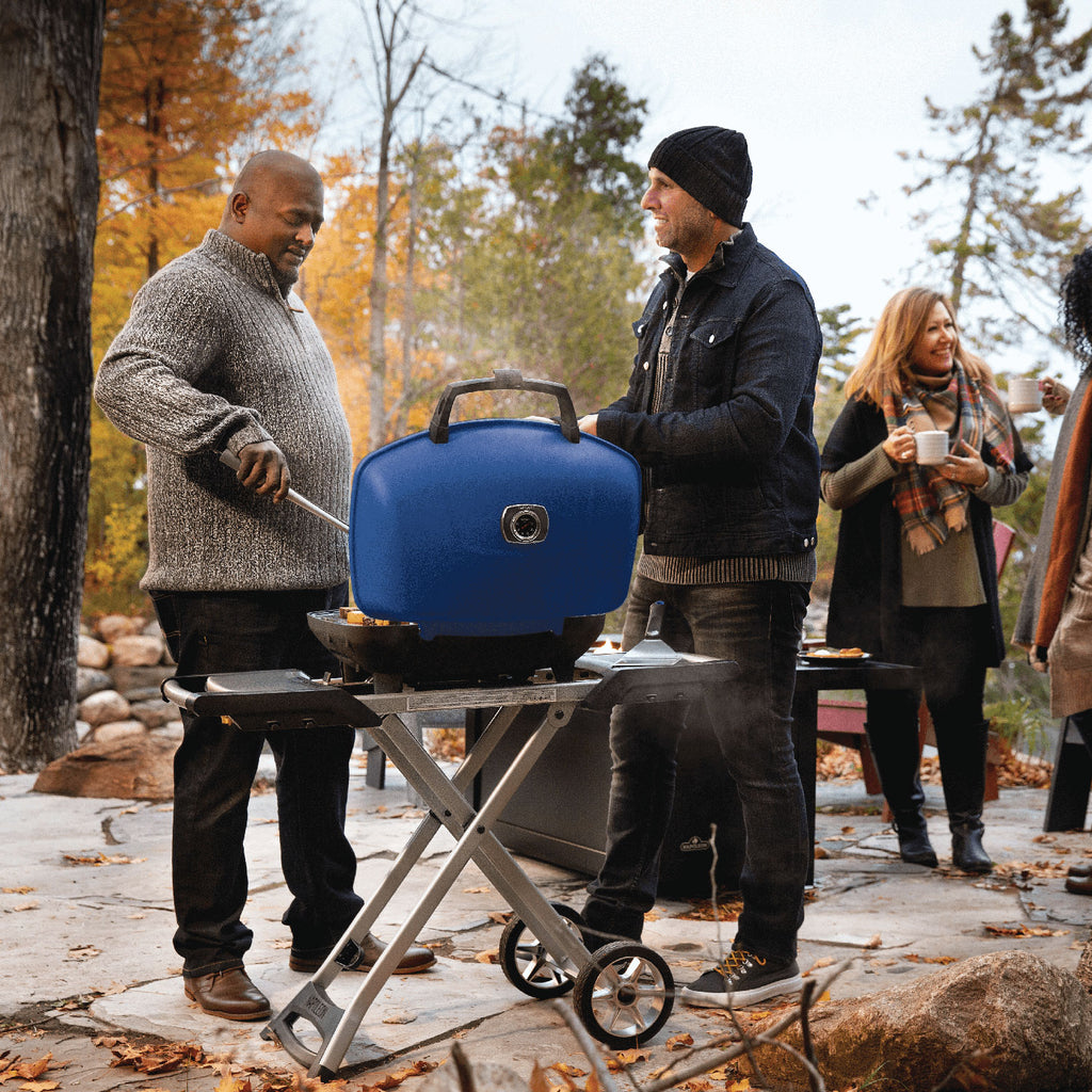 Portable propane grill with blue lid on scissor cart outdoors near seating