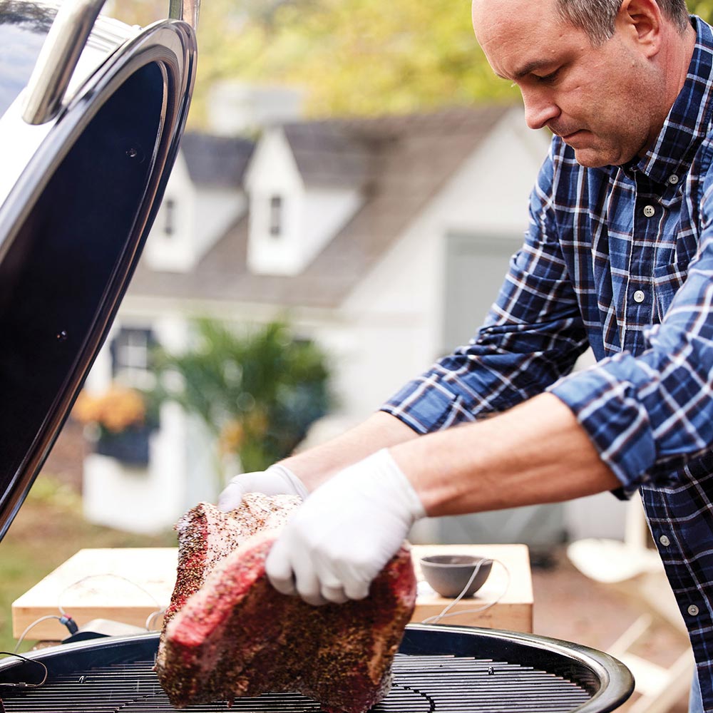 Weber 24-inch charcoal grill with open lid and gloved hands placing meat