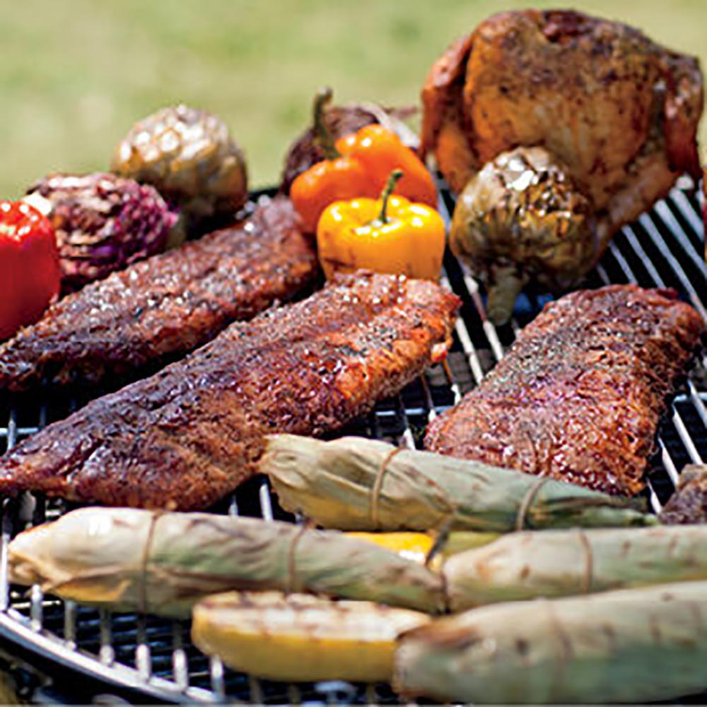 Charcoal kettle grill with grilled meats, sausages, and peppers on the cooking grate.