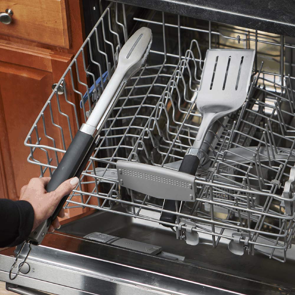 Stainless steel spatula and tongs with black handles in a dishwasher rack