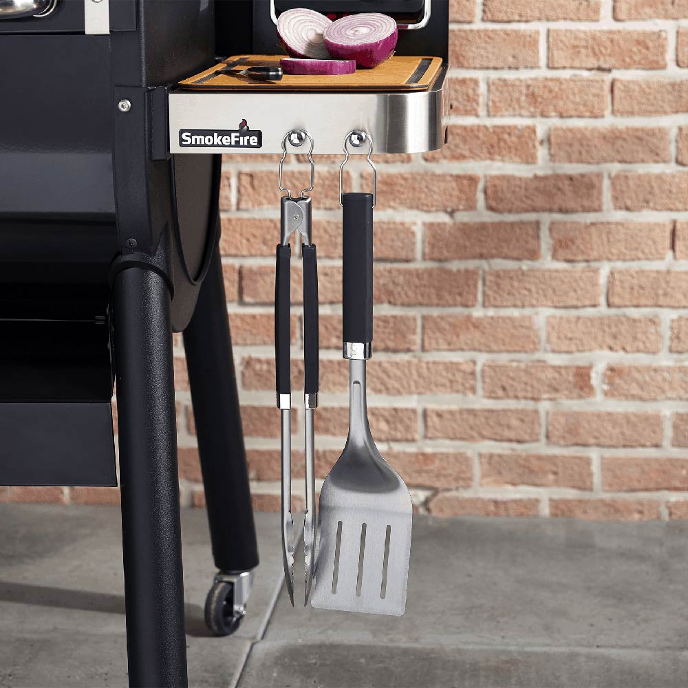 Stainless steel spatula and tongs with black handles hanging on a grill