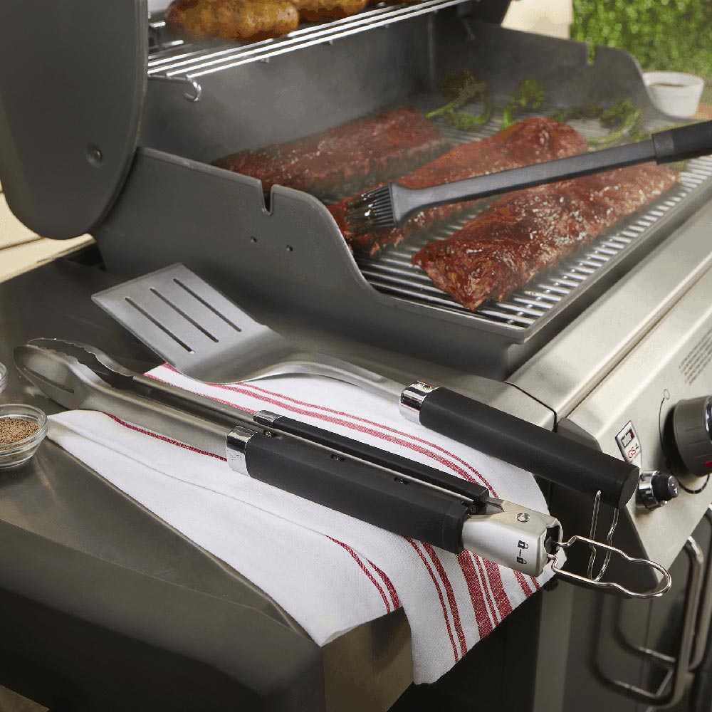 Black-handled grill tongs and stainless spatula beside cooked meat on grill grate