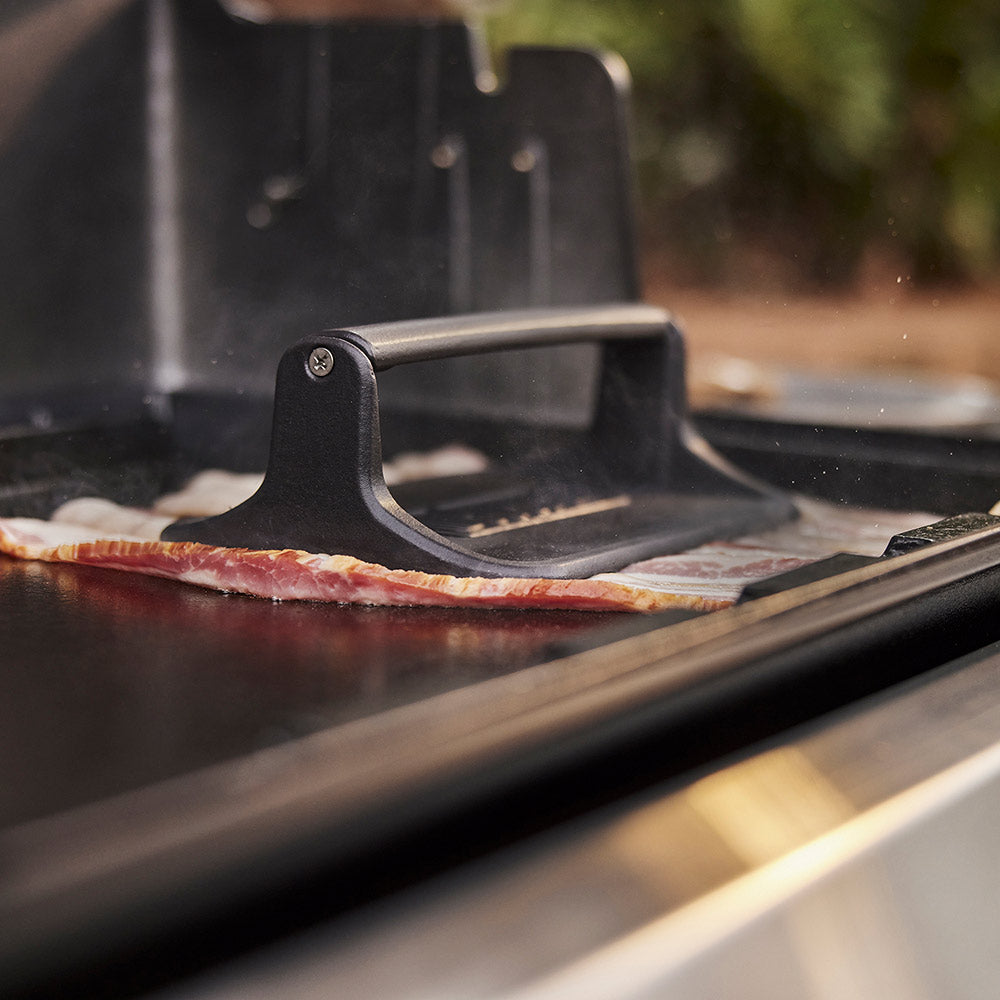 Griddle press pressing burgers on a grill, showing ridged plate