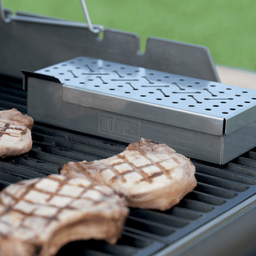 Weber smoker box with perforated lid on grill grates beside grilled meat