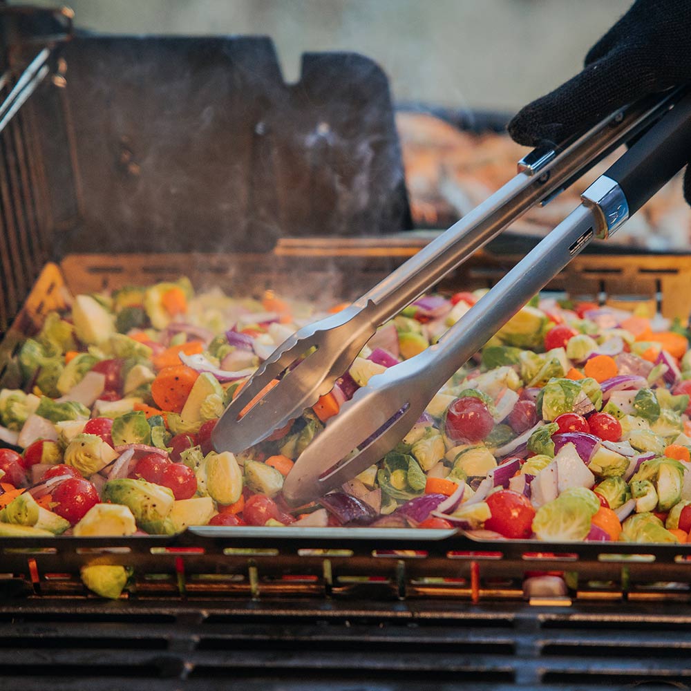 Roasting basket on a grill with tongs lifting food pieces