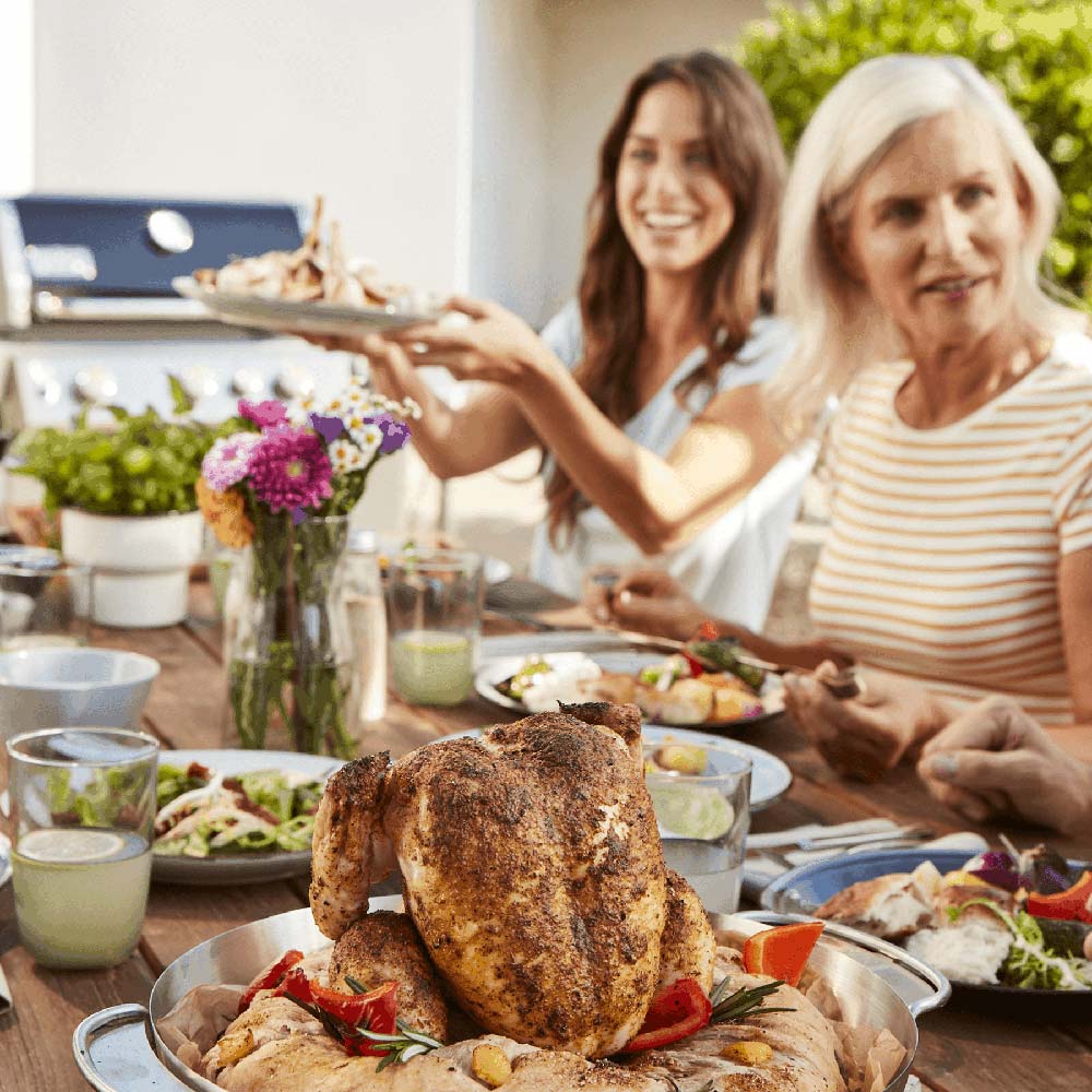 Whole chicken in stainless steel roaster basket on outdoor grill during meal