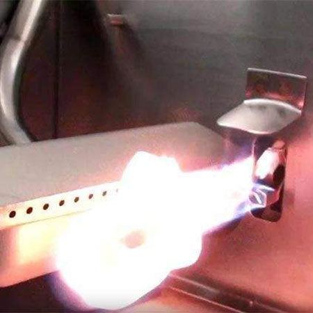 Lit gas grill burner under hood, bright flame and metal heat shield