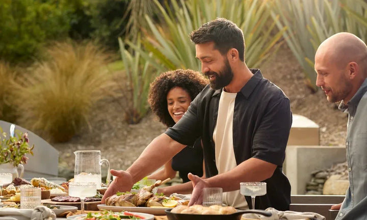 Friends and family gathered around an outdoor dining table loaded with grilled food from a Kickass Grills backyard setup